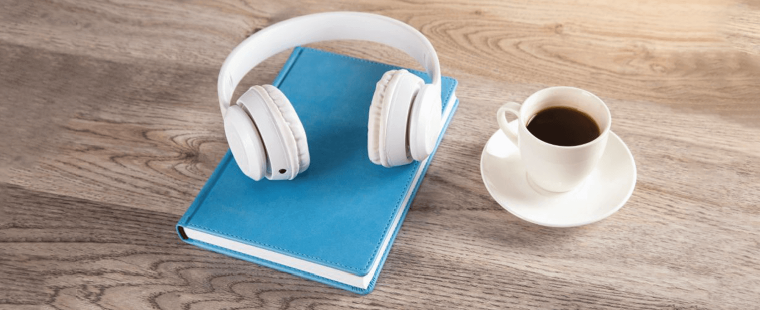 A cup of coffee, book and wireles headset on top of a desk.