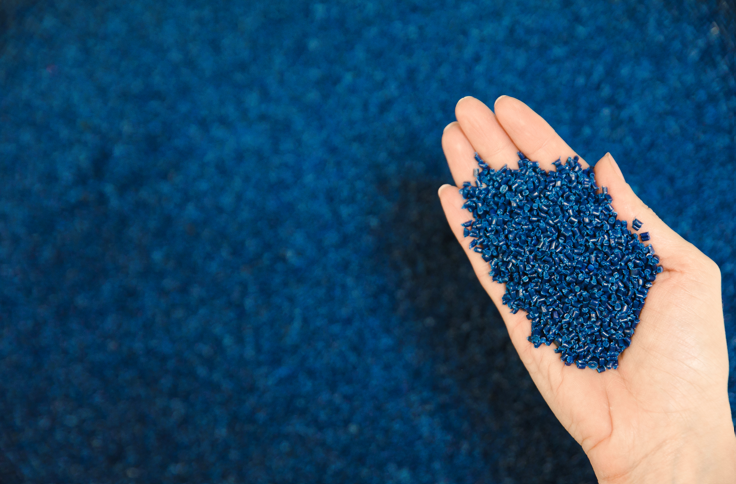 Female hand holding blue UBQ material pellets at her palm over a pile of pellets.